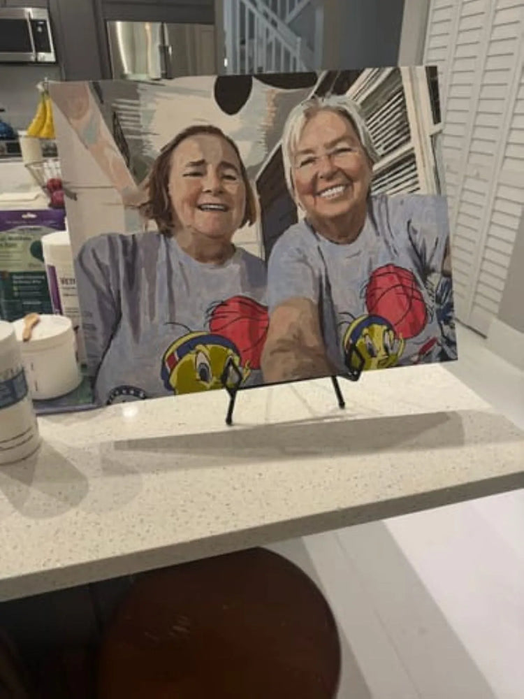 A printed photo print displayed on a small black stand on a kitchen counter. The photo shows two smiling women wearing matching gray T-shirts with a cartoon bird graphic and red basketballs.